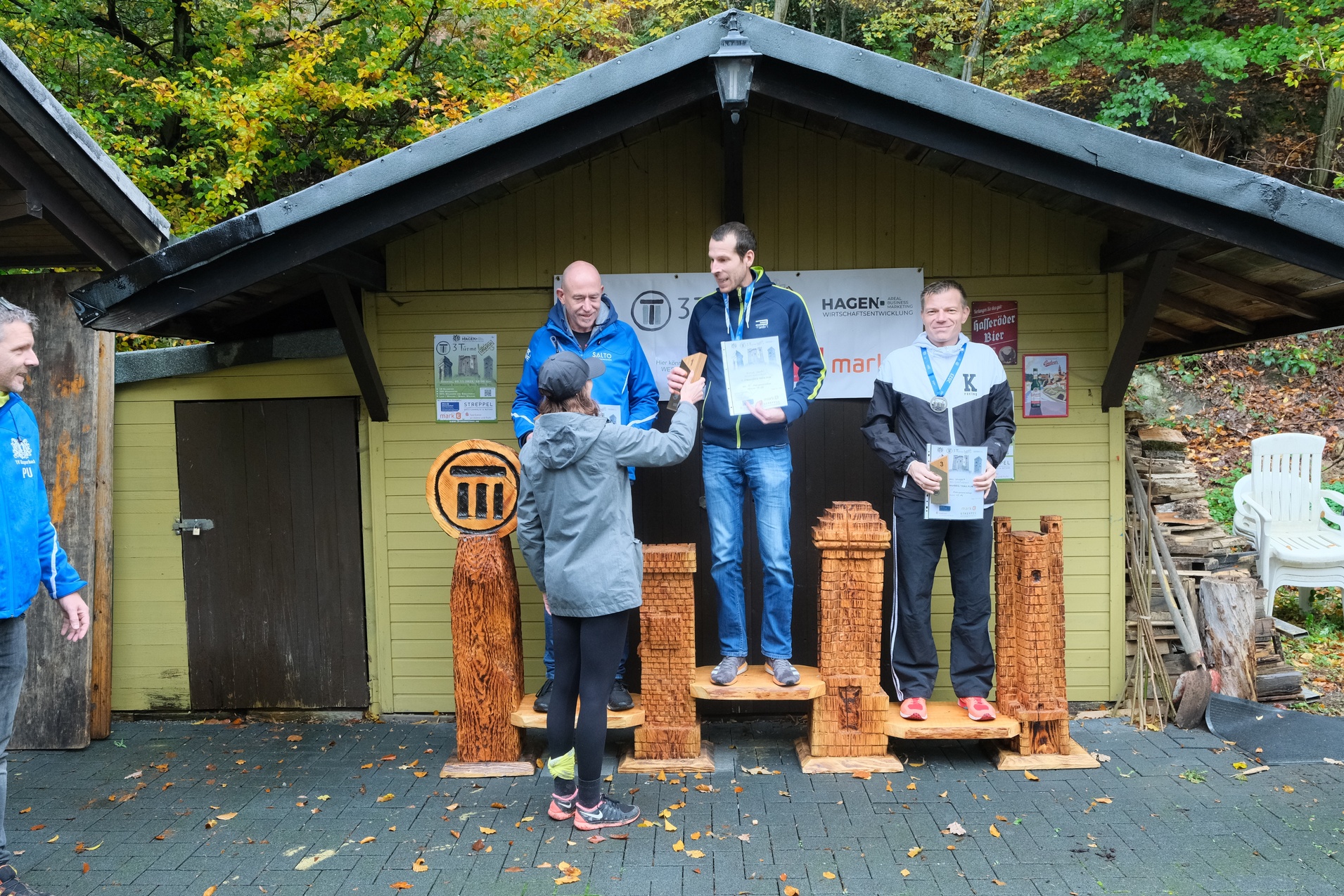 Siegerehrung mit Sportlern auf einem Podest vor einem Holzgeb&auml;ude, jeder h&auml;lt eine Urkunde in der Hand.