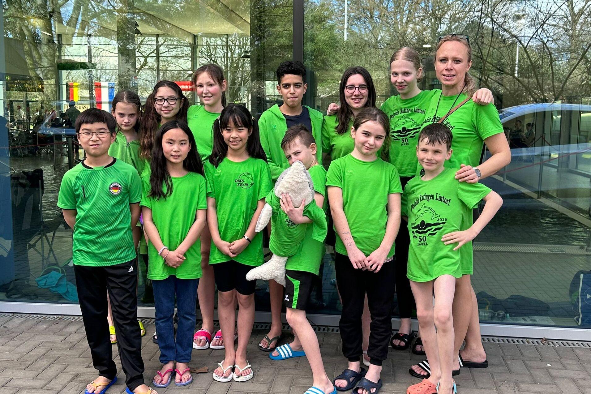 Gruppe von 14 Kindern und Jugendlichen in grünen T-Shirts, stehend vor großen Fenstern eines Schwimmbades.