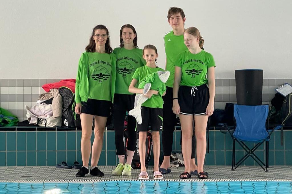 F&uuml;nf Schwimmer in gr&uuml;nen Shirts stehen l&auml;chelnd am Beckenrand eines Hallenbades.