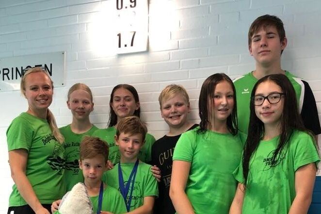 Gruppenfoto von acht Kinder und Jugendlichen in gr&uuml;nen T-Shirts, einem Trainer und einer Flipchart mit Sprungnoten im Hintergrund.