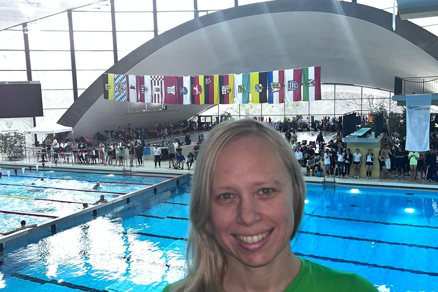 Frau mit blonden Haaren in grünem Shirt lächelt vor einem Schwimmbecken mit Zuschauern und Fahnen im Hintergrund.