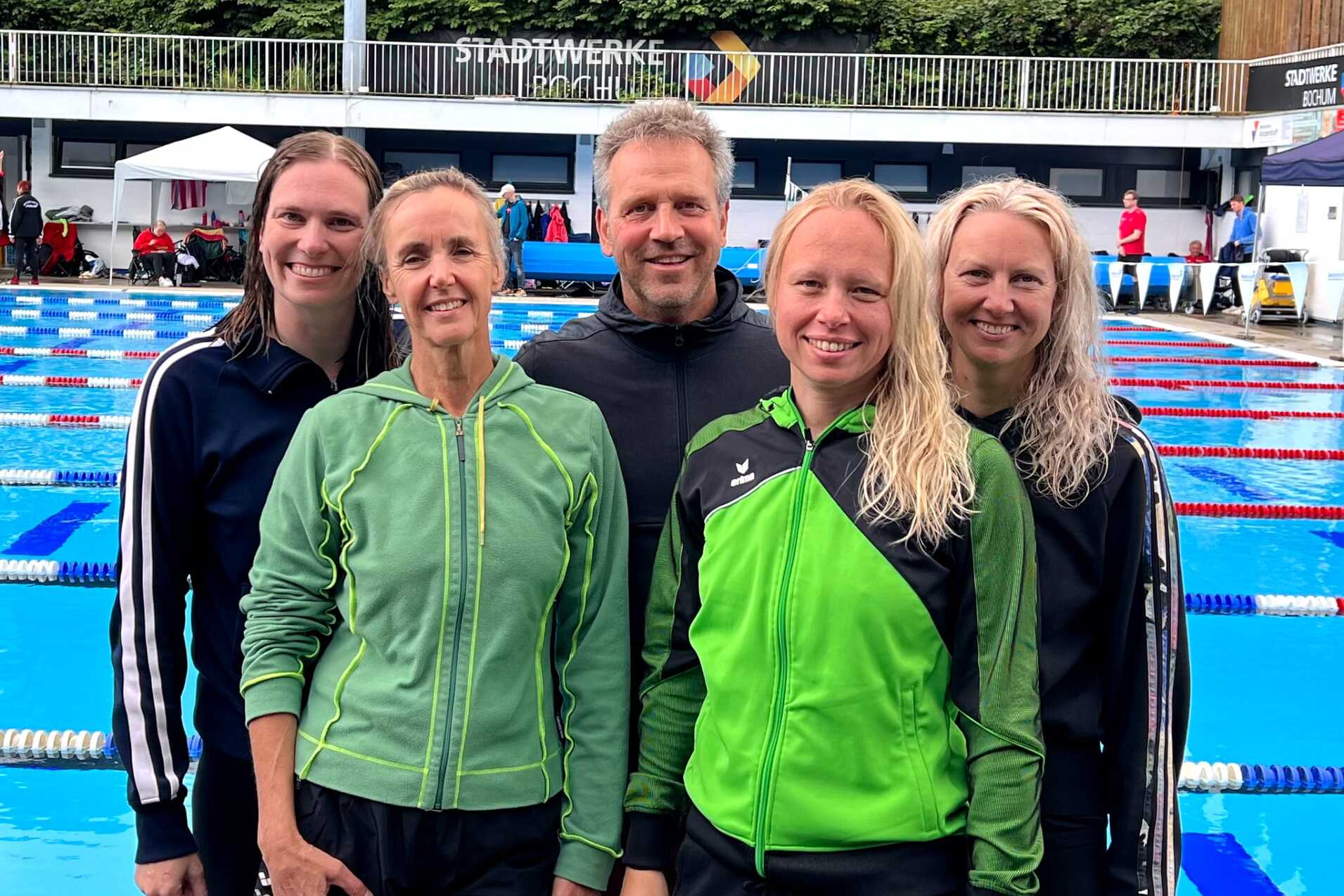 Fünf Schwimmtrainer stehen lächelnd am Beckenrand eines Freibades mit blauen Bahnen im Hintergrund.