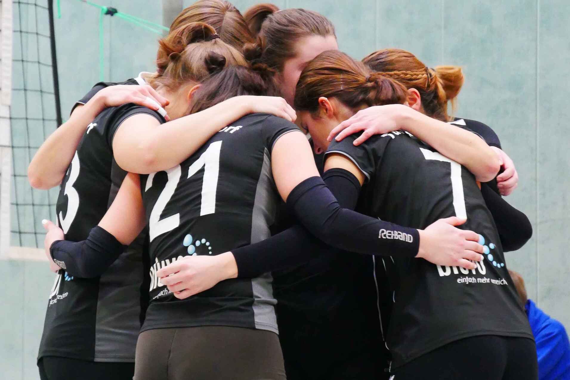 Sechs Volleyballspielerinnen umarmen sich in einer Sporthalle, alle in schwarzen Trikots und grünen Socken.