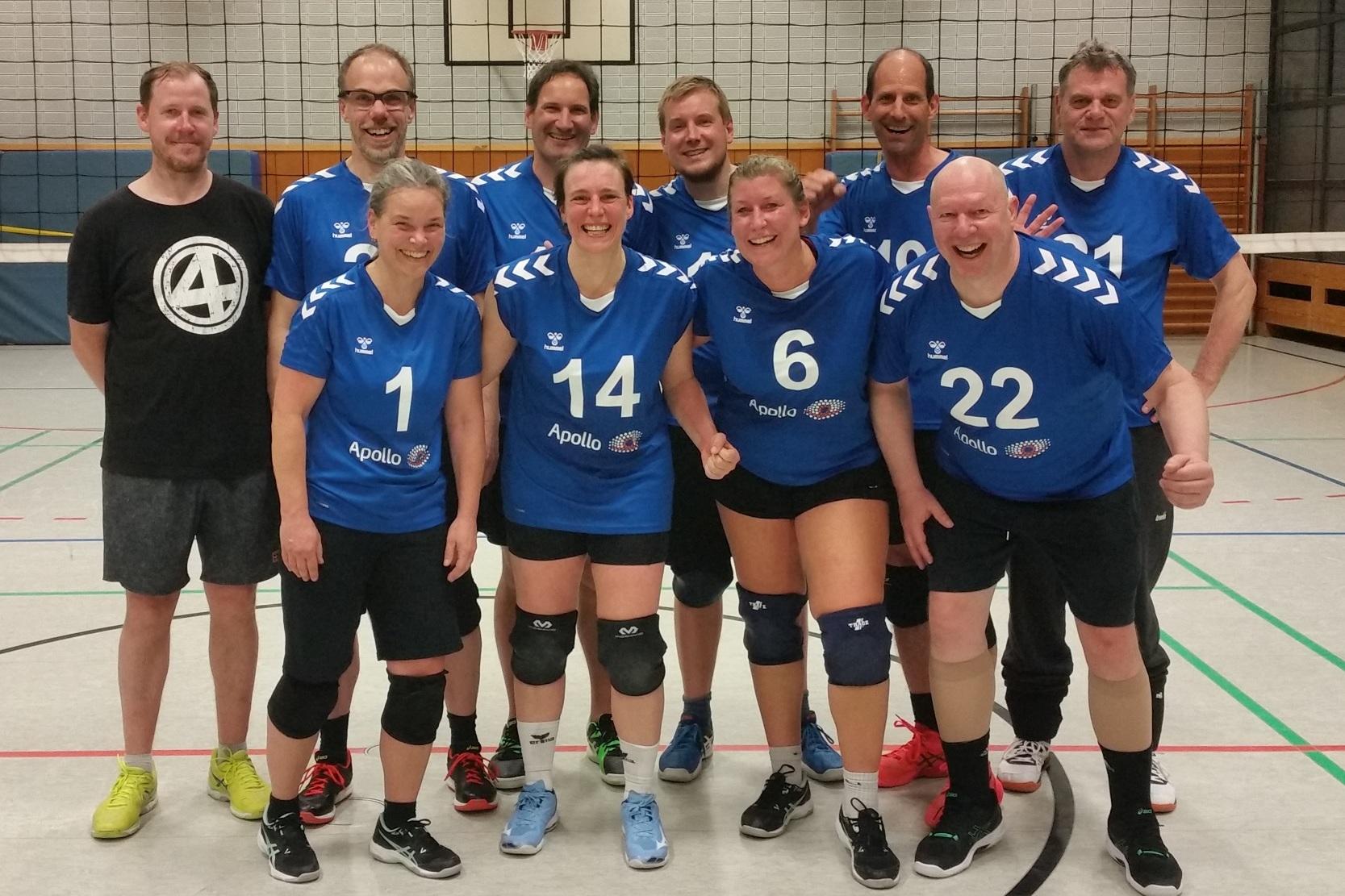 Gruppenfoto einer Volleyballmannschaft in blauen Trikots, lächelnd, auf einem Sportfeld mit Netz im Hintergrund.
