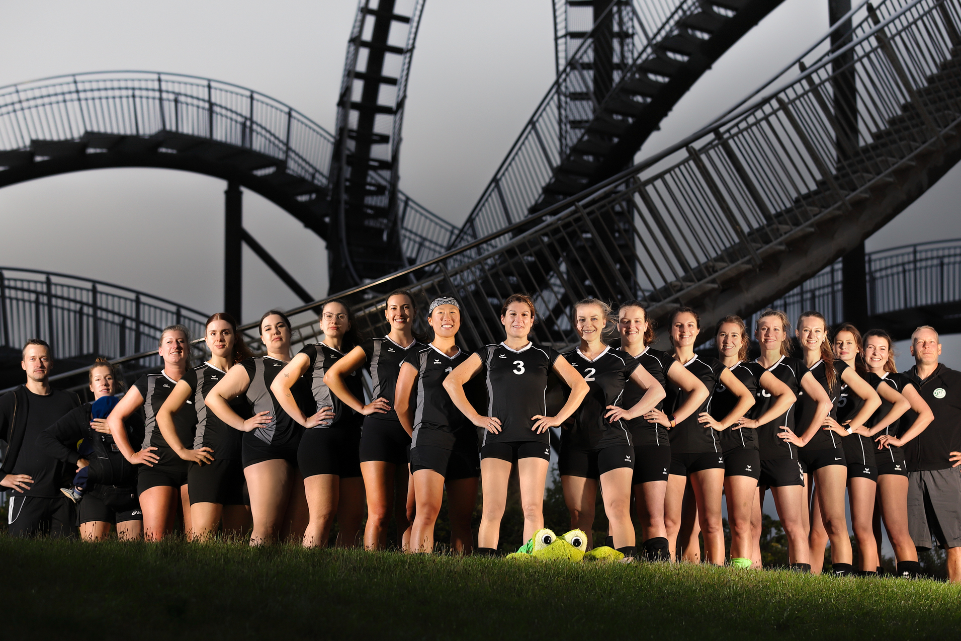 Gruppennaufnahme eines Damenvolleyballteams in Schwarz, posierend vor einer Achterbahn.