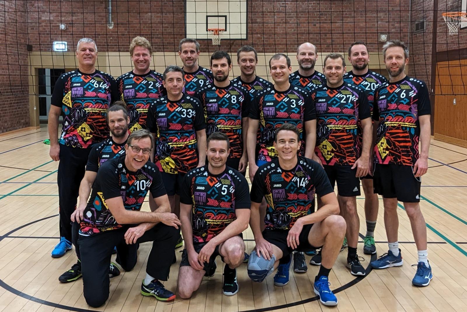 Gruppenfoto von 15 Männern in bunten Sporttrikots in einer Sporthalle, mit Volleyballnetz im Hintergrund.