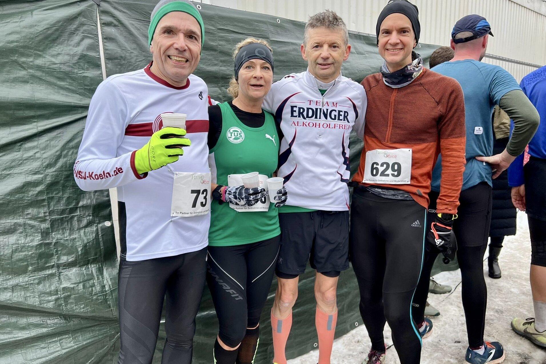 Vier Läufer posieren lächelnd vor einer grünen Plane, jeder mit einer Startnummer auf der Brust, im Winterwetter.