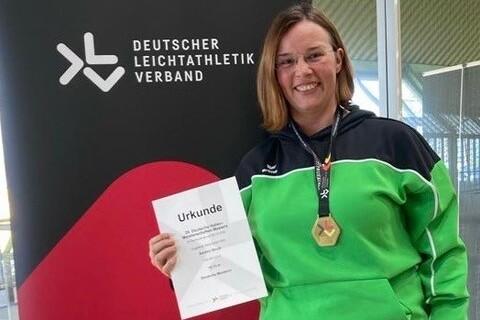 Frau in Sportjacke h&auml;lt Urkunde und Medaille vor Banner des Deutschen Leichtathletikverbands.