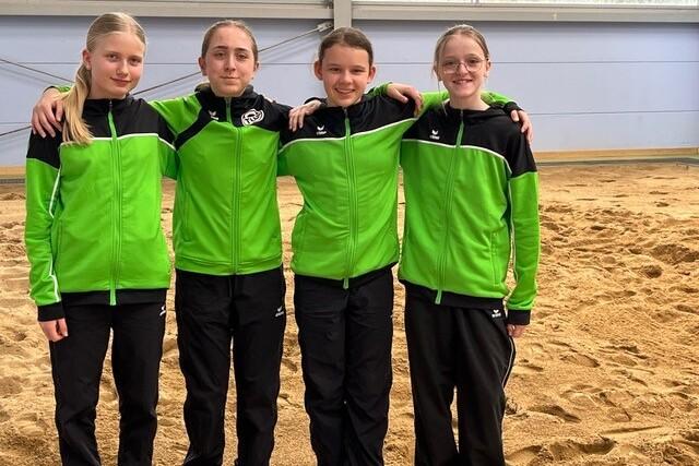 Vier Mädchen in grünen Sportjacken stehen arm in arm vor einer sandigen Fläche in einer Sporthalle.