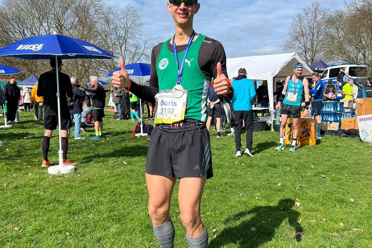 L&auml;ufer mit Startnummer 5192 posiert mit Daumen hoch auf einer Wiese, umgeben von Zuschauern und Veranstaltungspavillons.