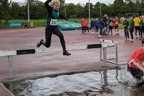 Sportliche Jugendliche springt w&auml;hrend eines Wettkampfs &uuml;ber eine H&uuml;rde, unter der sich eine Wasserpf&uuml;tze befindet.