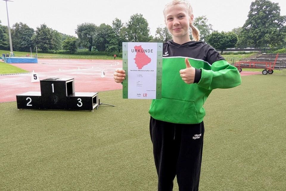 M&auml;dchen in Sportkleidung h&auml;lt stolz eine Urkunde und zeigt mit Daumen hoch, im Hintergrund eine Laufbahn und Podest.