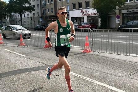 Läufer in Wettkampfbekleidung sprintet auf einer Straße entlang von Absperrgittern und Wohngebäuden.