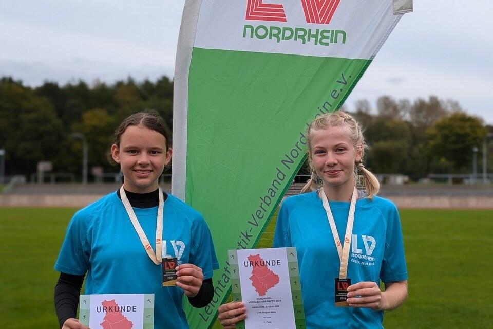 Zwei junge Athletinnen in blauen T-Shirts halten Urkunden und Medaillen vor einem Banner des LV Nordrhein.