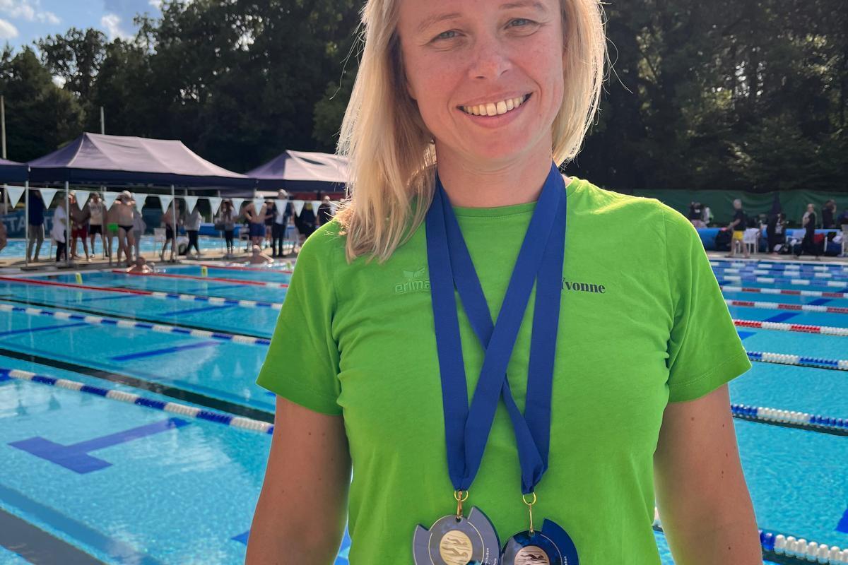 Sportlerin mit blonden Haaren, grüner T-Shirt, lächelnd, trägt zwei Medaillen um den Hals vor einem Schwimmbecken.