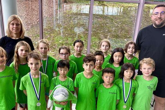 Gruppe von Kindern in gr&uuml;nen Shirts mit Medaillen und einem Ball, zusammen mit zwei Betreuern in einem Raum.