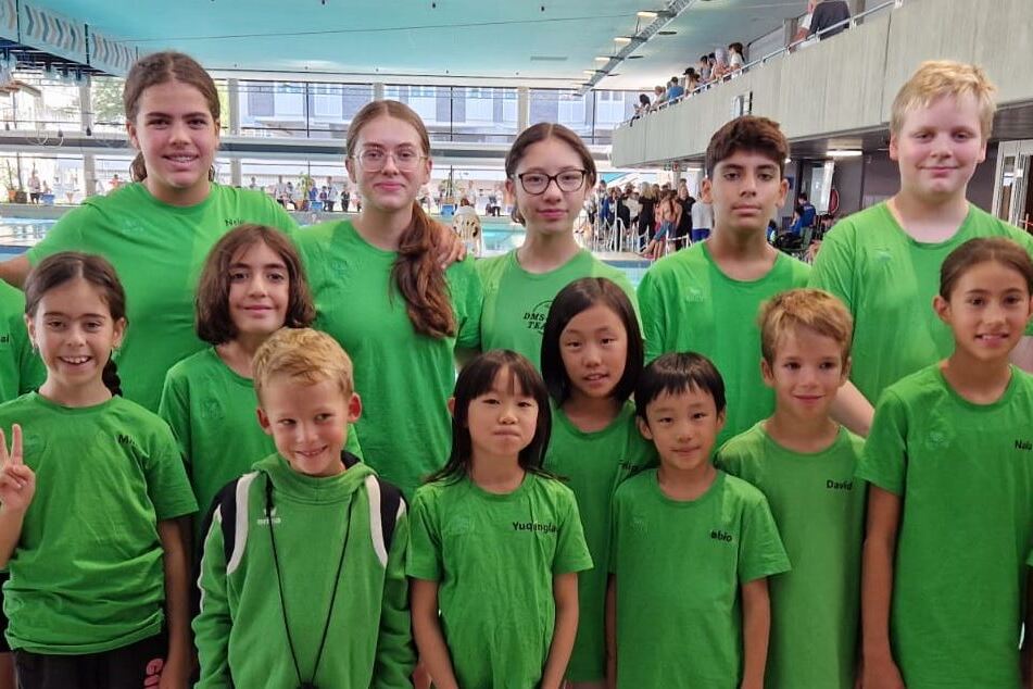 Gruppe von Schwimmern in gr&uuml;nen T-Shirts vor einem Schwimmbecken, l&auml;chelnd und posierend f&uuml;r ein Gruppenfoto.