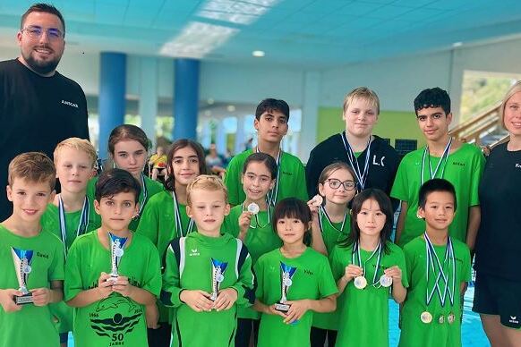 Gruppe junger Schwimmer in gr&uuml;nen Shirts mit Medaillen und Pokalen, posierend nach einem Wettkampf am Schwimmbad.