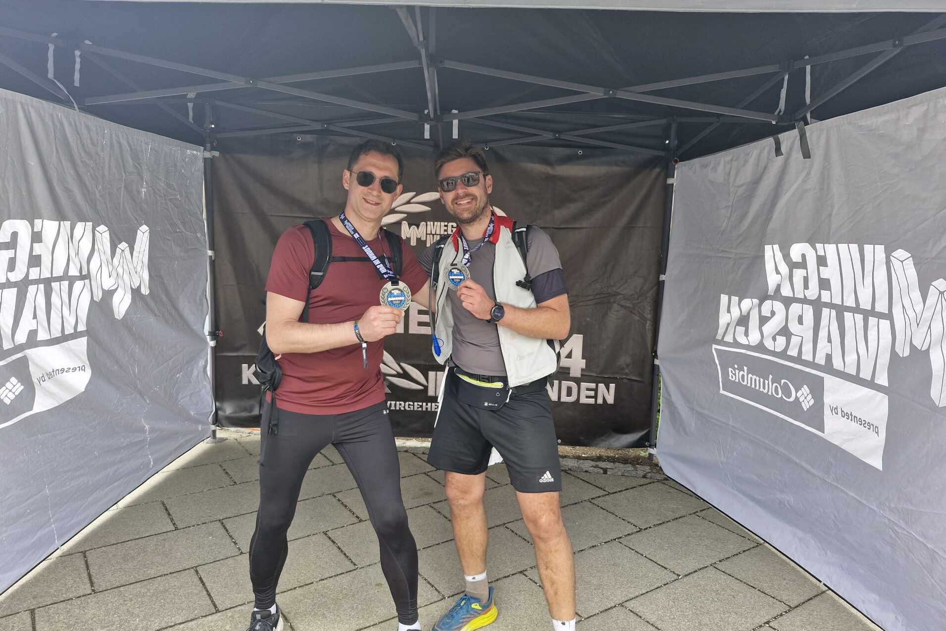 Zwei Sportler posieren unter einem Zeltdach und halten Medaillen in den Händen. Sie tragen Sportkleidung und Rucksäcke.