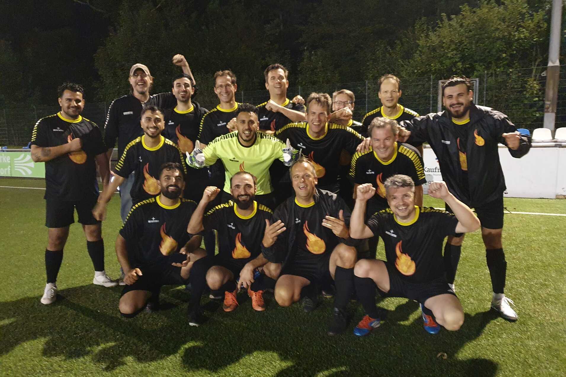 Gruppenfoto von zw&ouml;lf Fu&szlig;ballspielern in schwarzen Trikots mit gelben Details, posierend auf einem Kunstrasenplatz.