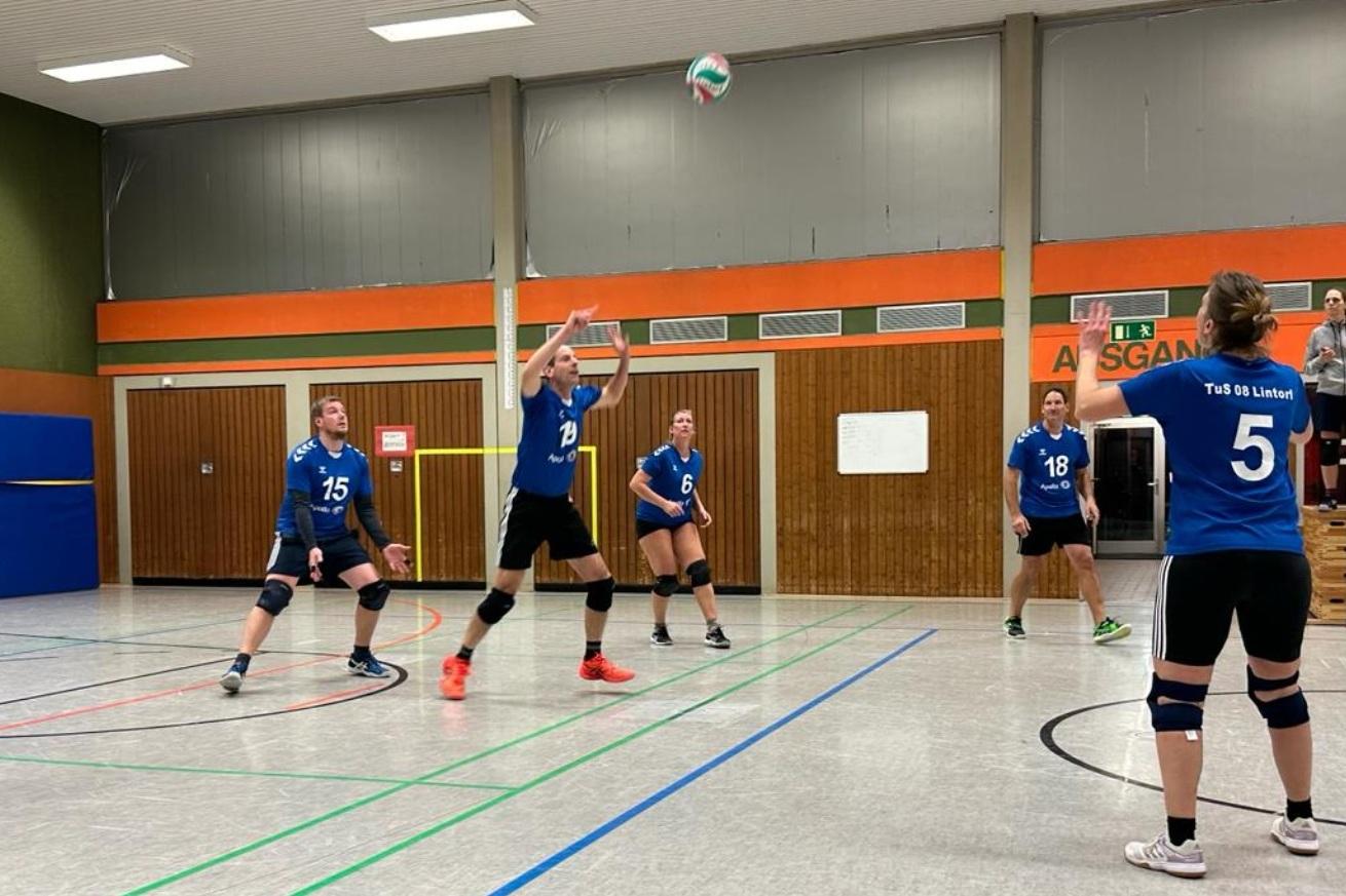 Sechs Volleyballspielerinnen in blauen Trikots in einer Halle während eines Spiels; eine Spielerin hebt den Ball an.