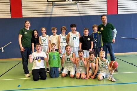 Gruppe von Jungen in Basketballtrikots steht l&auml;chelnd auf einem Sportplatz, umgeben von Trainern. Basketball in der Mitte.