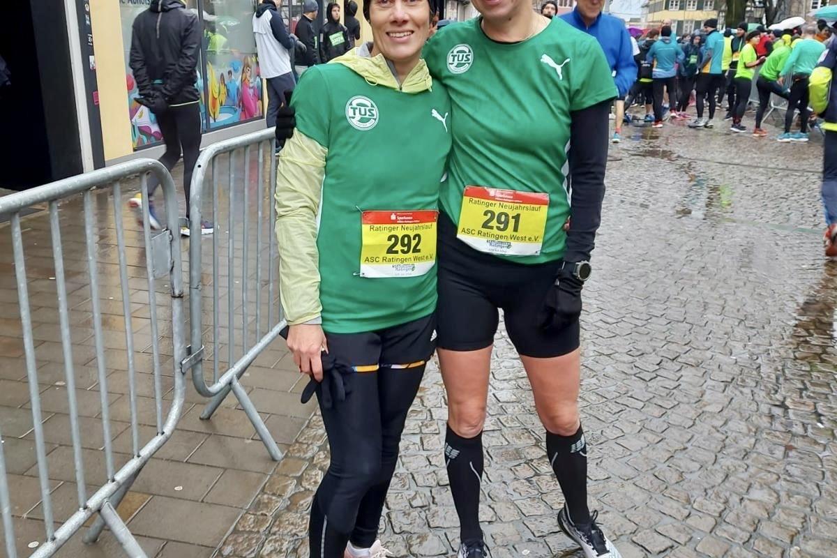 Zwei Frauen in grünen Laufshirts mit Startnummern stehen auf einer nassen Straße vor einer Laufveranstaltung.