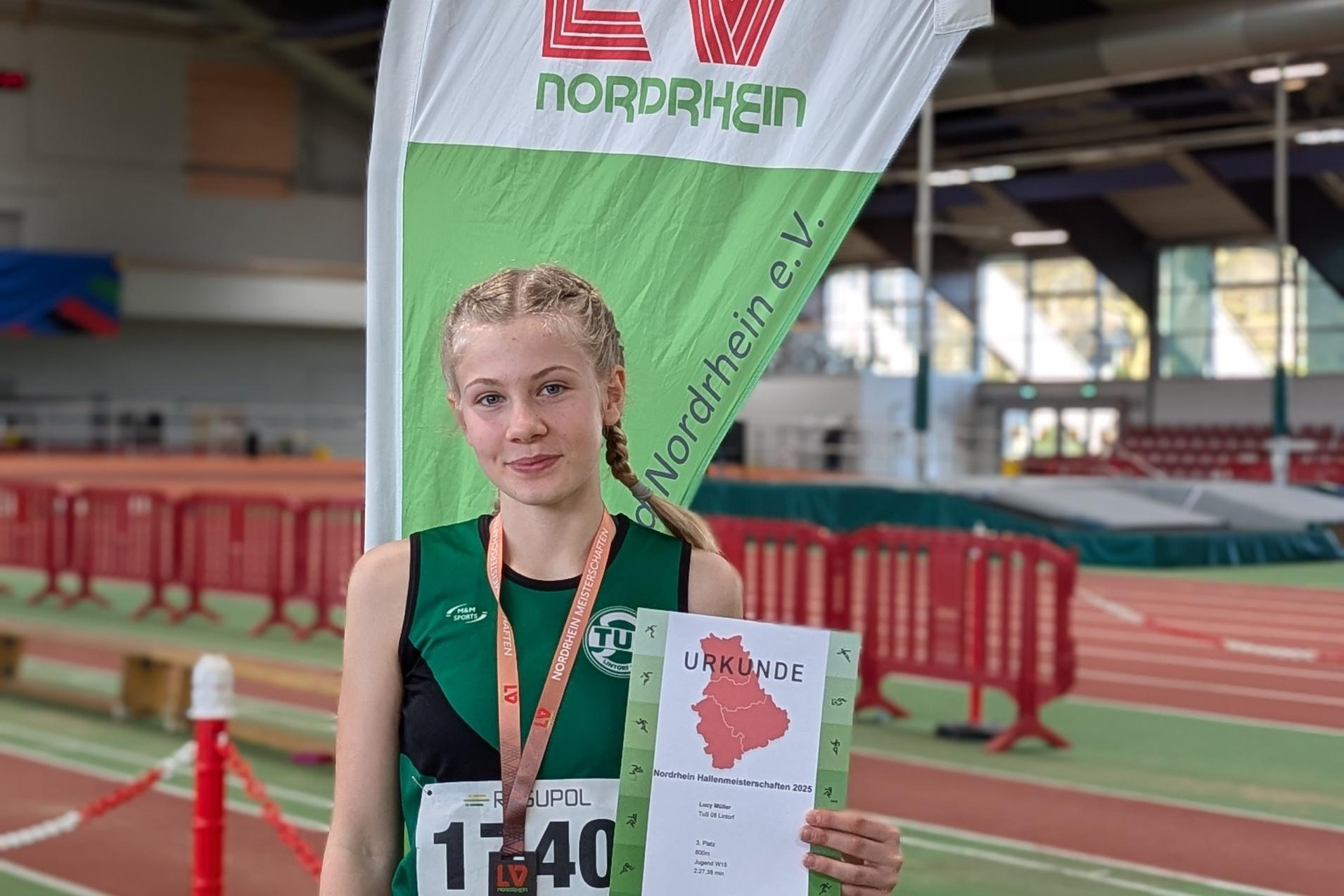Mädchen in Sportkleidung hält Urkunde und Medaille vor einem Banner mit dem Logo des LV Nordrhein e.V. in einer Sporthalle.