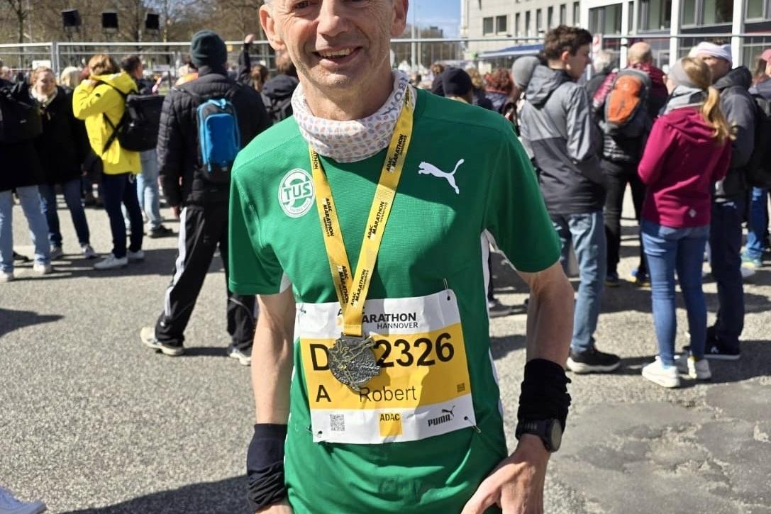 Mann in gr&uuml;ner Sportbekleidung steht mit Medaille nach dem Marathon unter blauem Himmel im Gedr&auml;nge von Zuschauern.