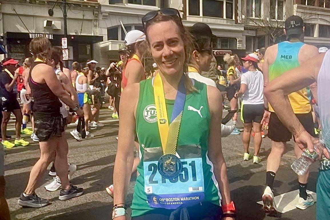 L&auml;uferin l&auml;chelt mit Finisher-Medaille nach dem Boston Marathon, umgeben von weiteren Teilnehmern und Stadtarchitektur.