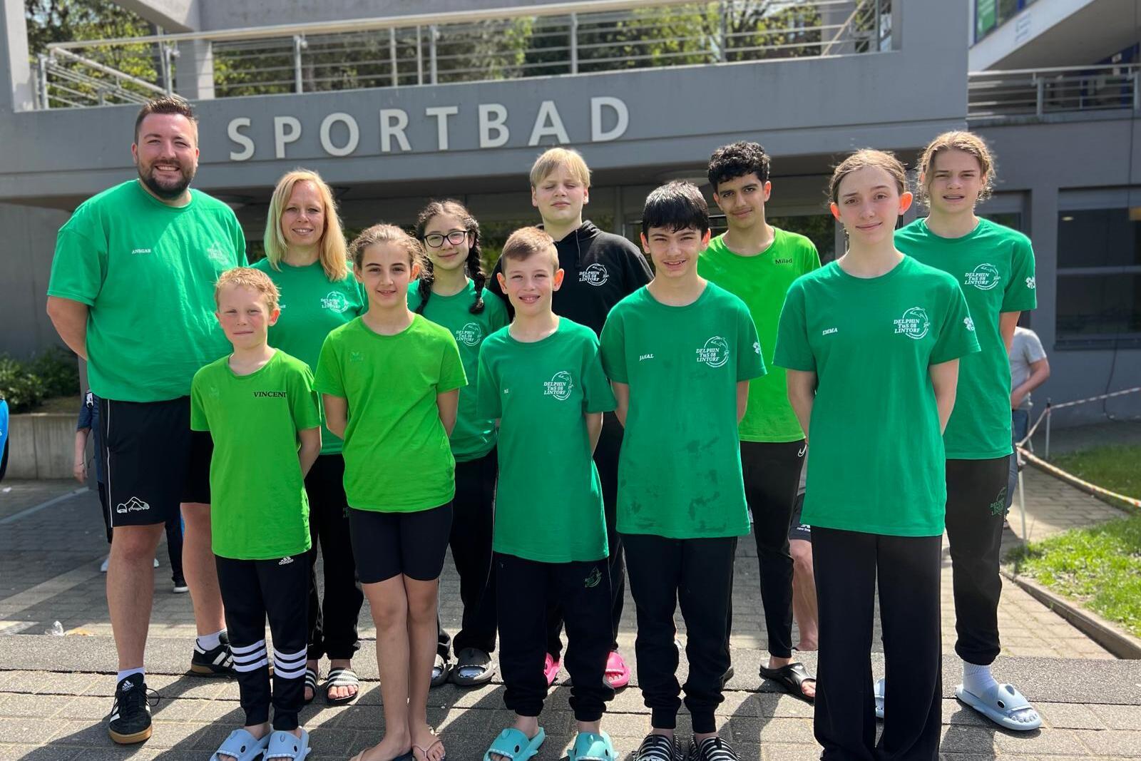 Gruppe von Schwimmern und Trainern in gr&uuml;nen T-Shirts vor dem Sportbad, l&auml;chelnd auf einem Gehweg.