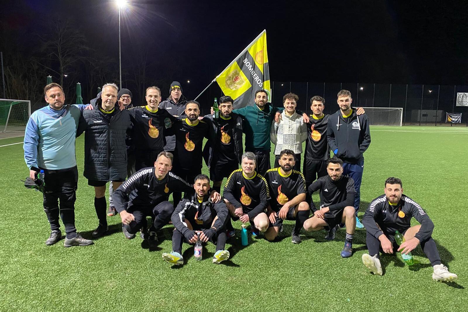 Eine Gruppe von 15 Fußballspielern in schwarzen Trikots posiert mit einer Fahne auf einem Kunstrasenplatz bei Nacht.