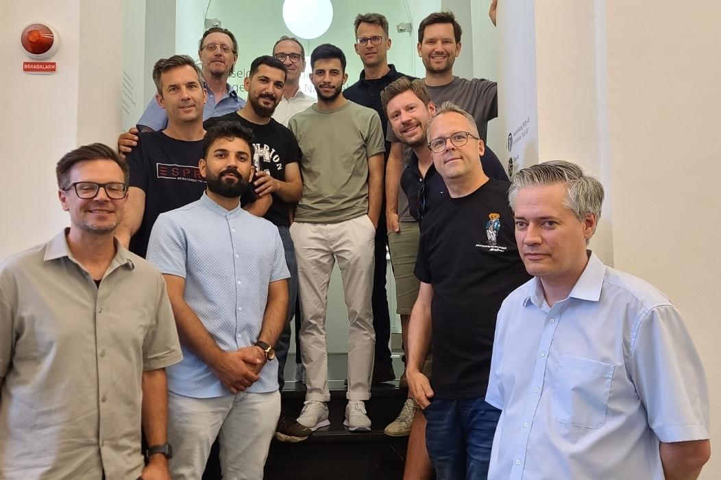 Gruppenfoto von zwölf Männern, die auf einer Treppe stehen, mit modernen Innenräumen und Deckenleuchten im Hintergrund.