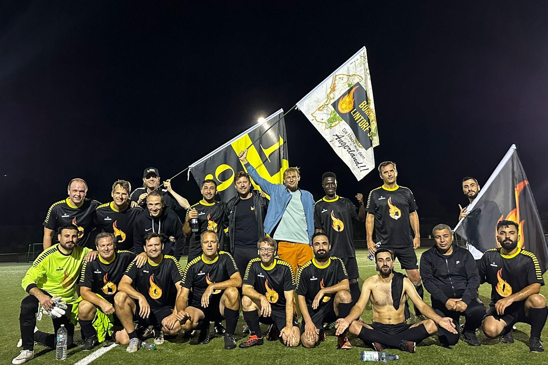 Mannschaft von 20 Fußballspielern in schwarzer Kleidung posiert mit Fahnen auf einem Kunstrasenplatz bei Nacht.
