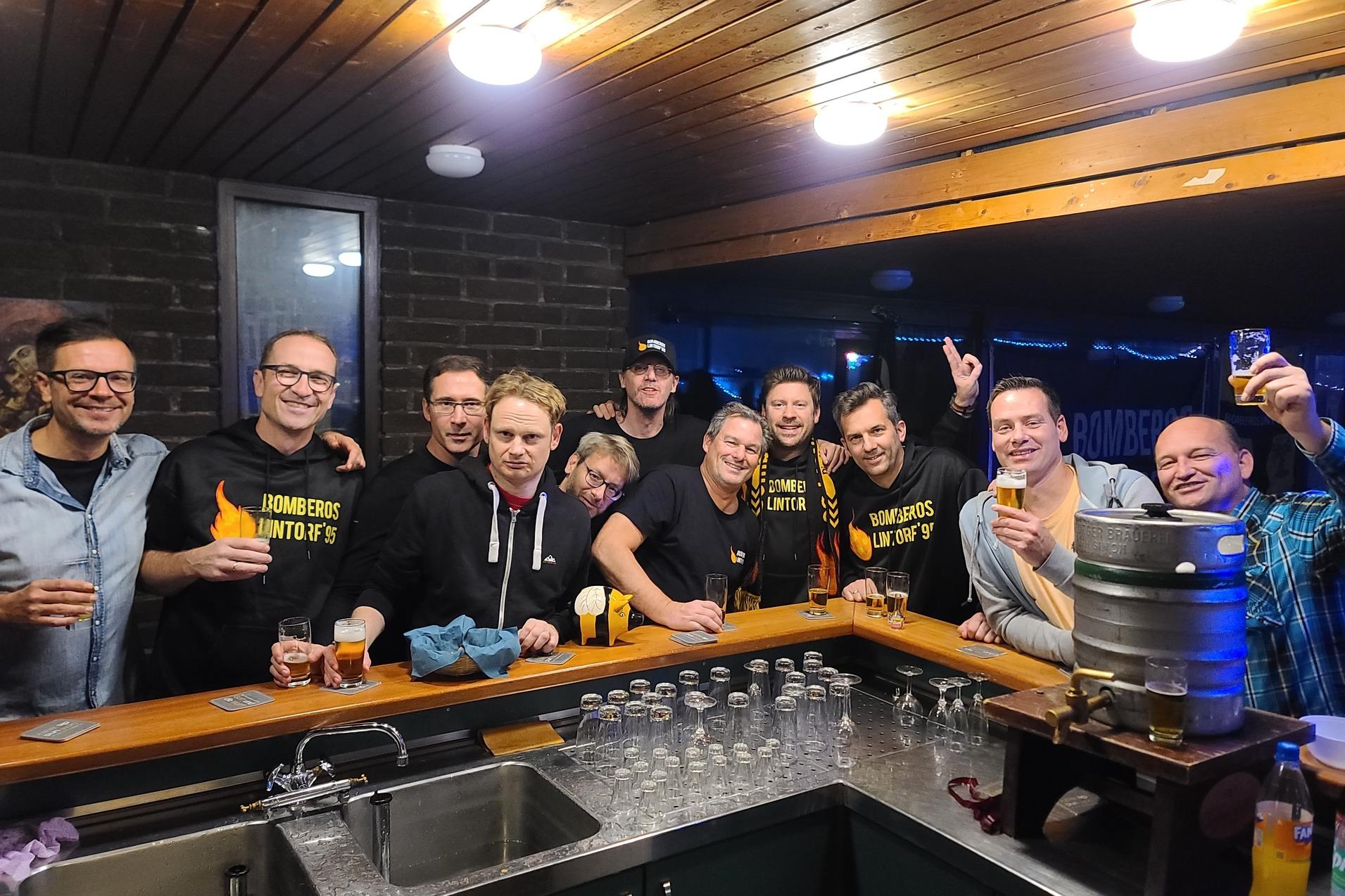 Gruppe von zw&ouml;lf Personen l&auml;chelt an einer Bar, h&auml;lt Gl&auml;ser und posiert vor einem Bierfass.