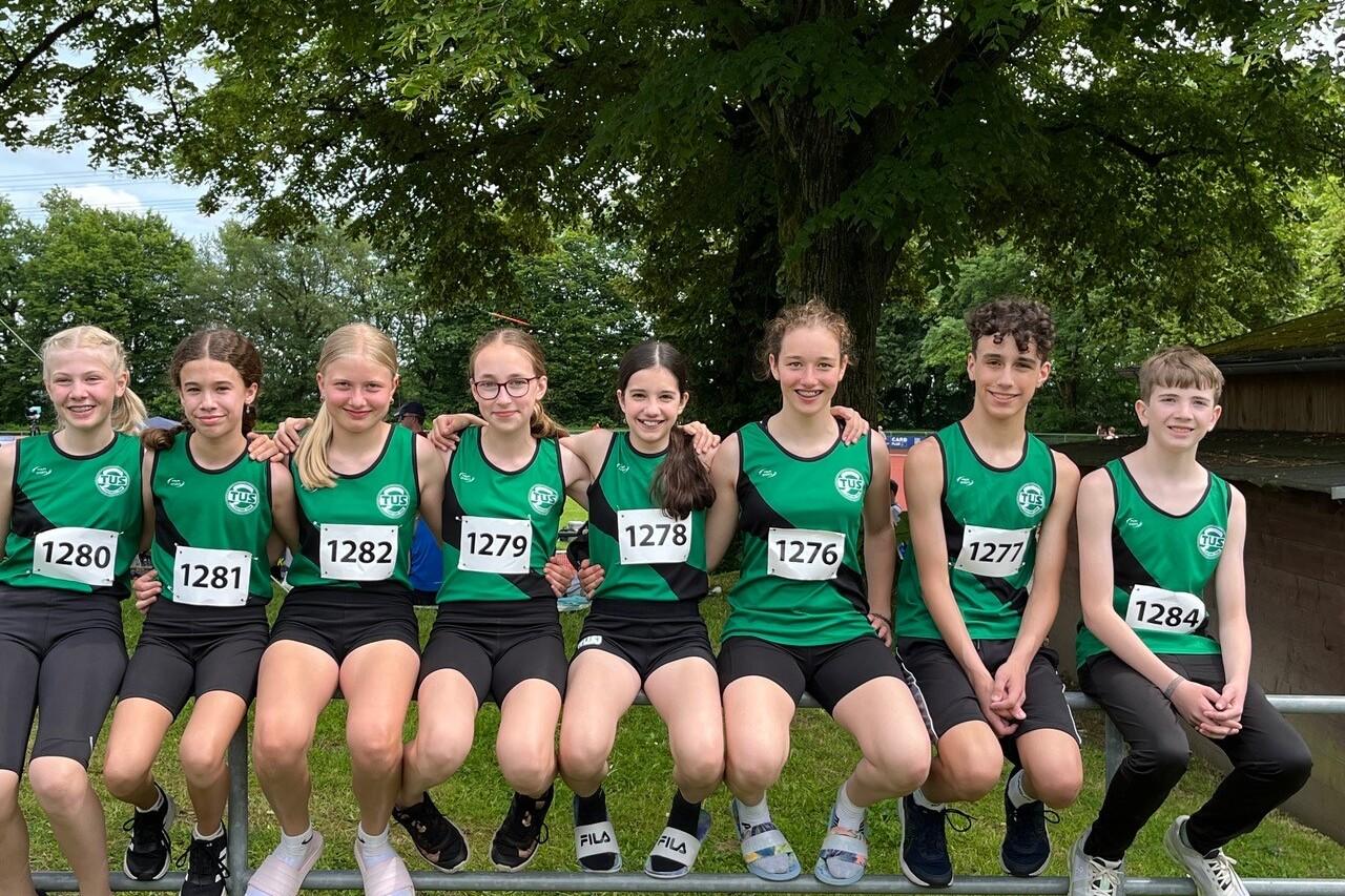 Gruppe von acht jungen Athleten in grünen Trikots sitzt lächelnd auf einem Zaun, Rennnummern sichtbar.