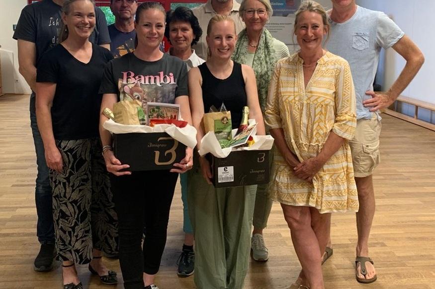 Gruppenfoto von zehn Personen, zwei halten Geschenkkörbe, lächelnd in einem Raum mit Holzfußboden.