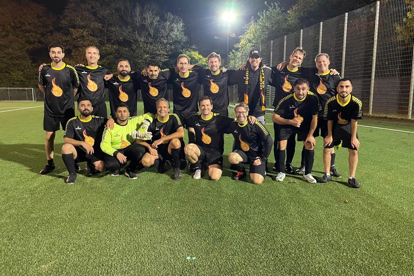 Gruppe von 14 Fußballspielern in schwarzen Trikots mit orangefarbenem Logo posieren gemeinsam auf einem Kunstrasenplatz.