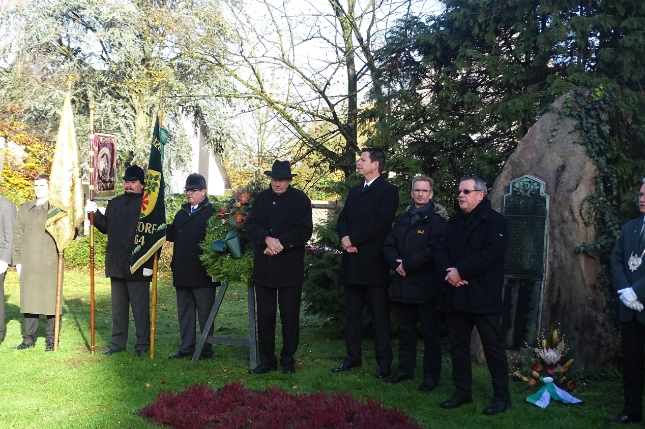 Gruppen von Menschen, in dunkler Kleidung, stehen um einen Gedenkstein und halten Fahnen und einen Kranz.