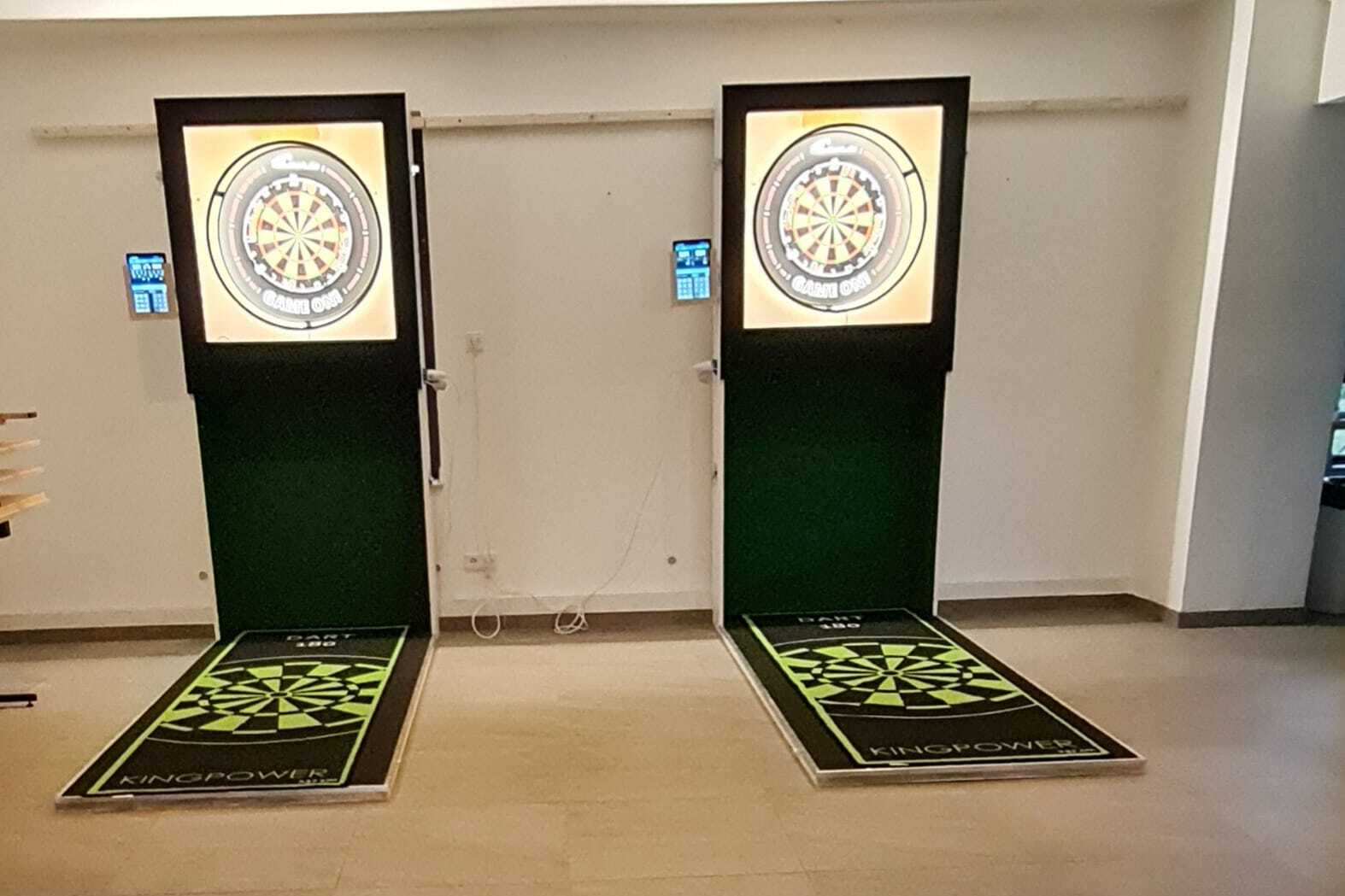 Zwei elektronische Dartscheiben mit farbigen Anzeigen in einem modernen Raum, davor grüne Wurfbereiche.