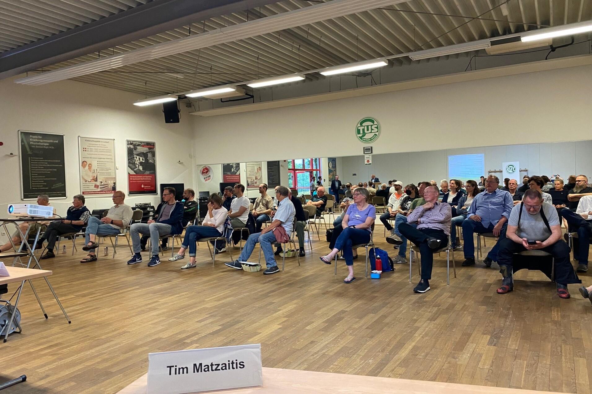 Zusammenkunft von Personen in einem Raum, die auf St&uuml;hlen sitzen und einem Vortrag folgen. Tische im Vordergrund.