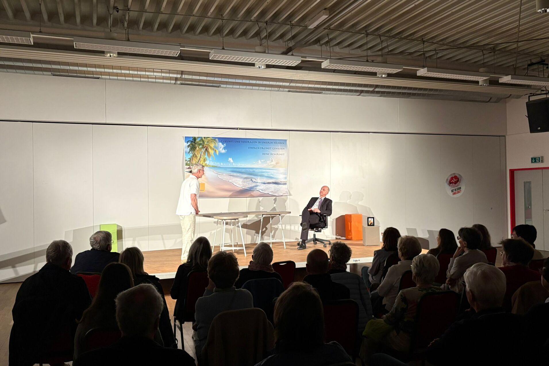 Zwei Personen auf einer B&uuml;hne, einer spricht, w&auml;hrend Publikum in Stuhlreihen zuh&ouml;rt. Hintergrund mit Strandmotiv.