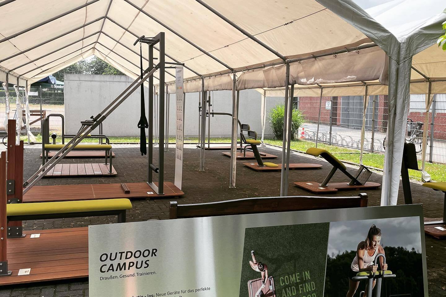Sportgeräte unter einem Zeltdach auf einem Outdoor-Campus mit einem Informationsschild von Dr. WOLFF im Vordergrund.