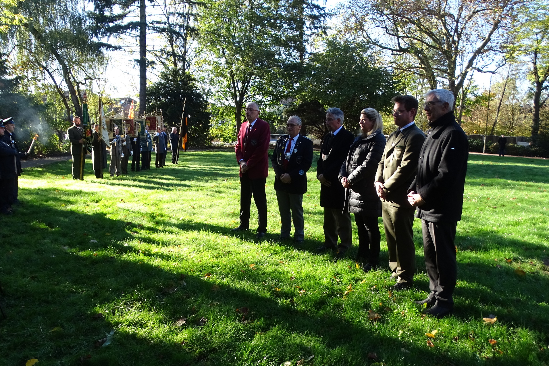 Gruppenbild von f&uuml;nf Personen, die an einer Gedenkfeier im Freien teilnehmen; im Hintergrund eine Ehrenwache.