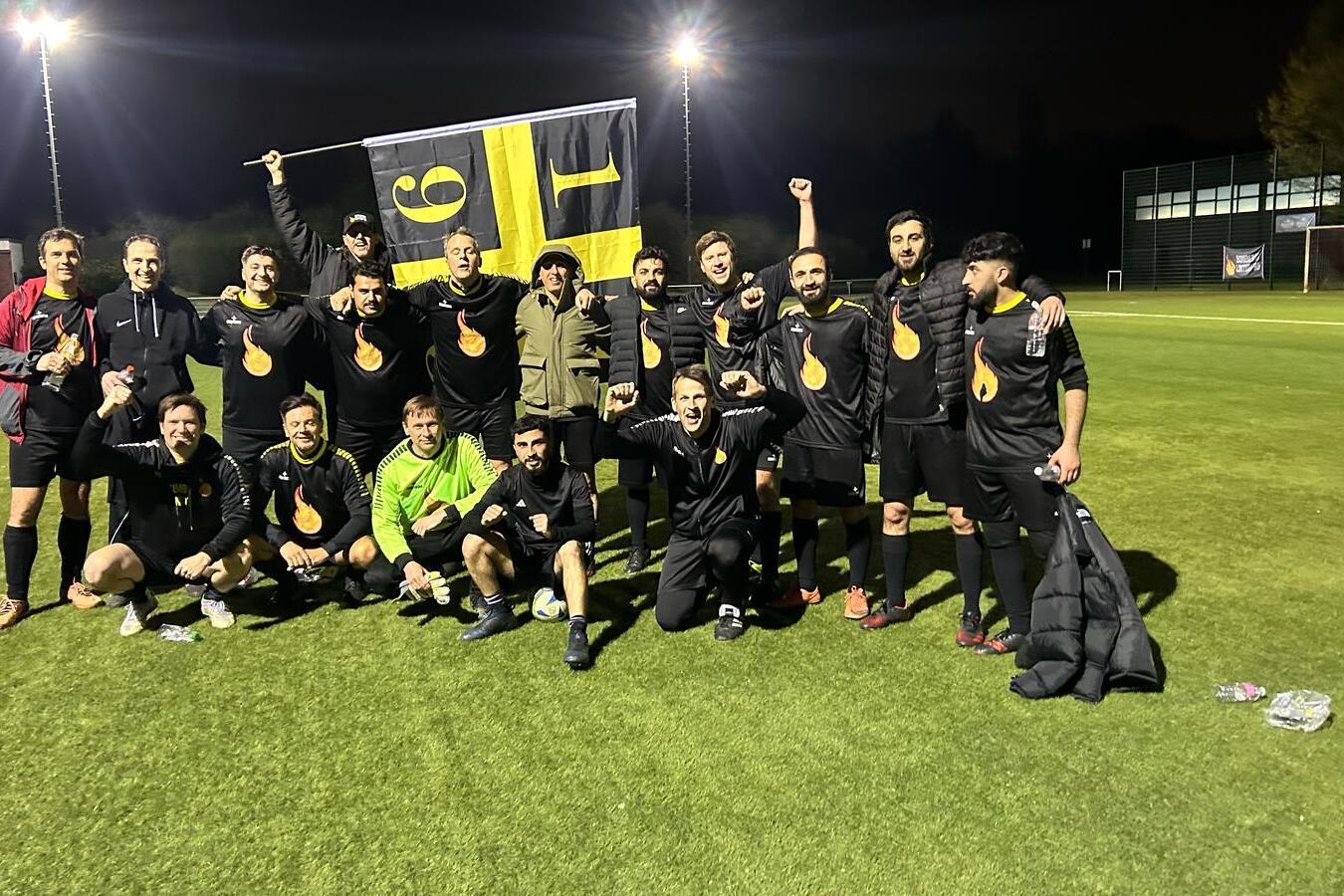 Feiernde Fu&szlig;ballmannschaft in schwarzen Trikots posiert mit einer Flagge auf dem Sportplatz bei Nacht.
