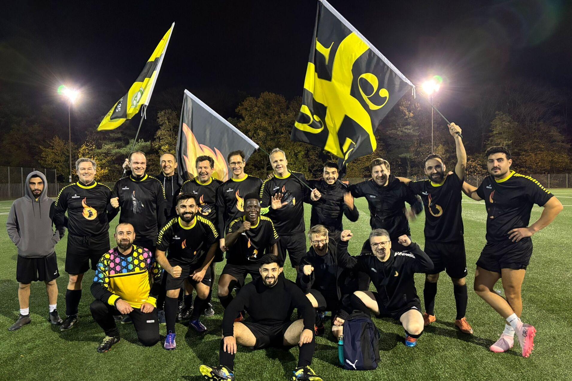 Gruppe von 17 Fußballspielern in schwarzen Trikots mit Flaggen auf einem Sportplatz bei Nacht.