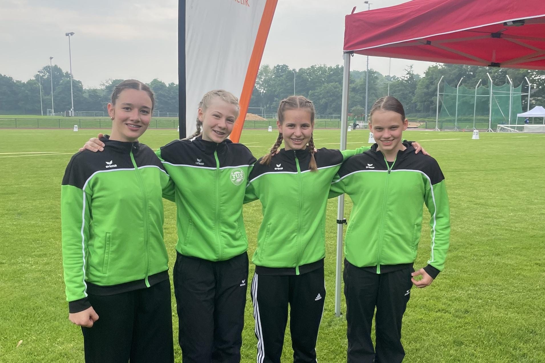 Vier M&auml;dchen in gr&uuml;nen Sportjacken posieren l&auml;chelnd vor einem Zelt und Banner der DJK Novesia auf einem Rasenplatz.
