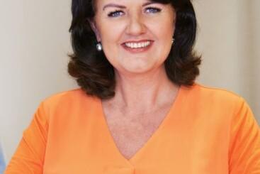 Portrait einer lächelnden Frau mit dunklen Haaren in einem orangefarbenen Oberteil, sitzend vor einem neutralen Hintergrund.