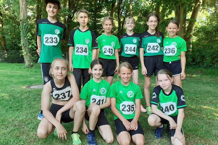 Gruppe junger Athleten in Sportkleidung mit Startnummern, auf einer Wiese im Freien, umgeben von B&auml;umen.