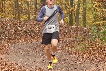 L&auml;ufer in Sportkleidung joggt auf einem mit Laub bedeckten Waldweg in einem herbstlichen Wald.